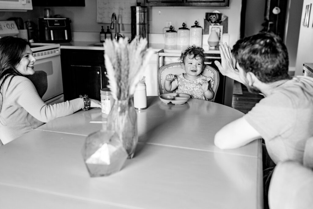 The dad and little girl are sitting at the kitchen table doing a high five with mom.