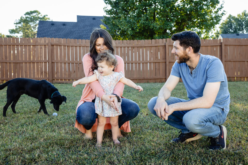 Mom, dad and little girl in the backyard. The little girl is snuggled between her mom's arms as they are all crotched on the ground. The family dog is also in the background with her ball.